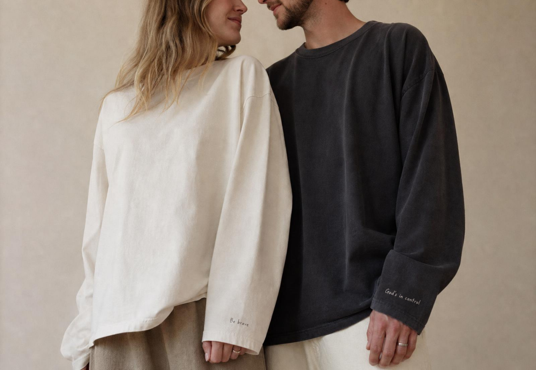 A couple in bone and charcoal long-sleeve tees; her sleeve reads 'be brave' and his reads 'God's in control'